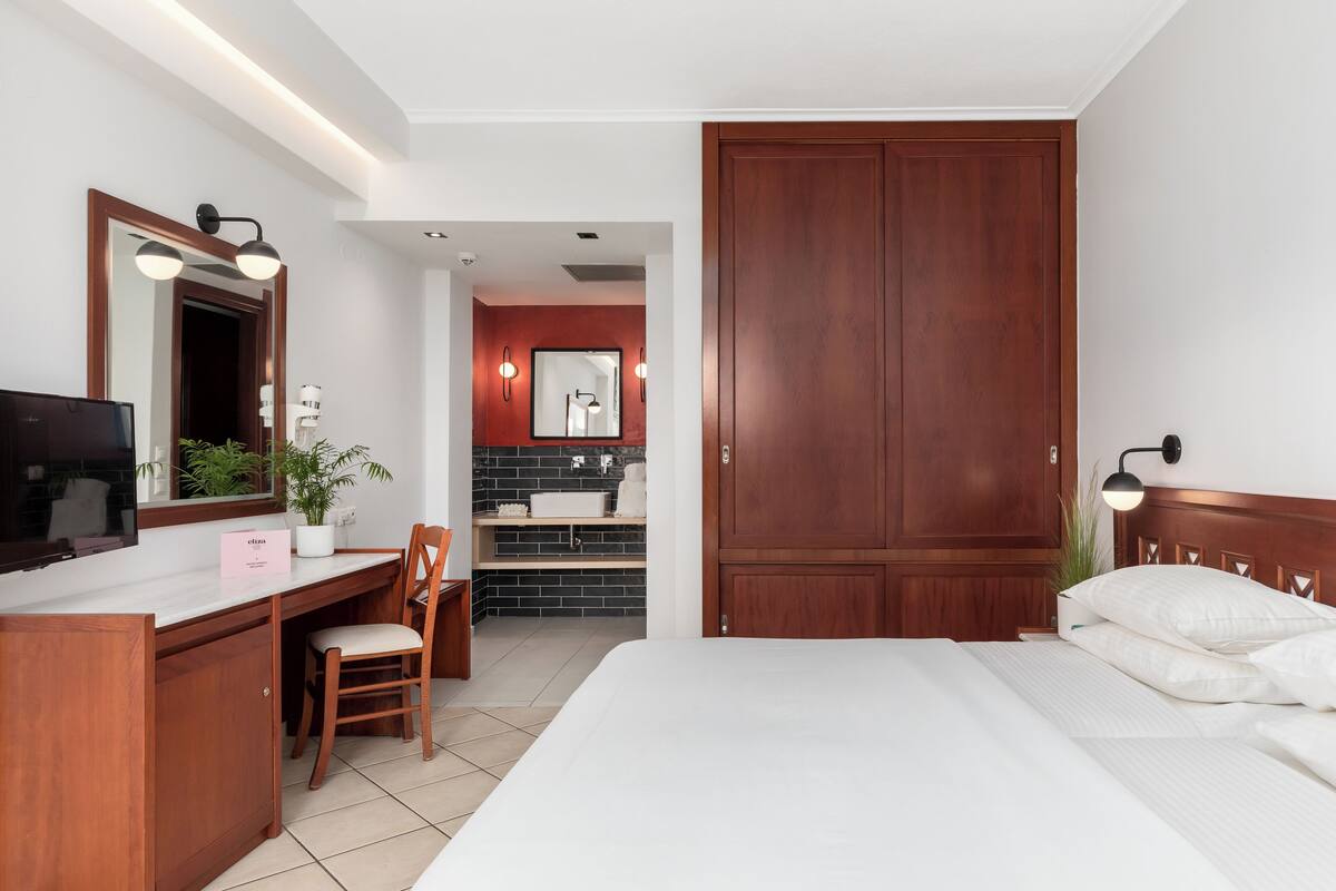 A hotel room with a bed, a desk with a chair, a television, a mirror, a wardrobe, and a bathroom visible through an open door.