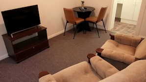 A living room with a flat-screen TV on a wooden stand, two beige armchairs, a small round table with a potted plant, and a dining area with two chairs. - Millicent Motel (Millicent)