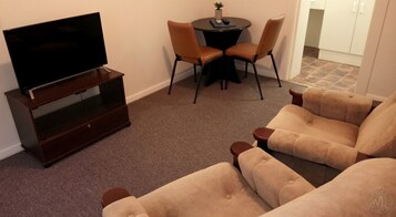 Una sala de estar con un televisor de pantalla plana sobre un soporte de madera, dos sillones color beige, una mesita redonda con una planta en maceta y un comedor con dos sillas.