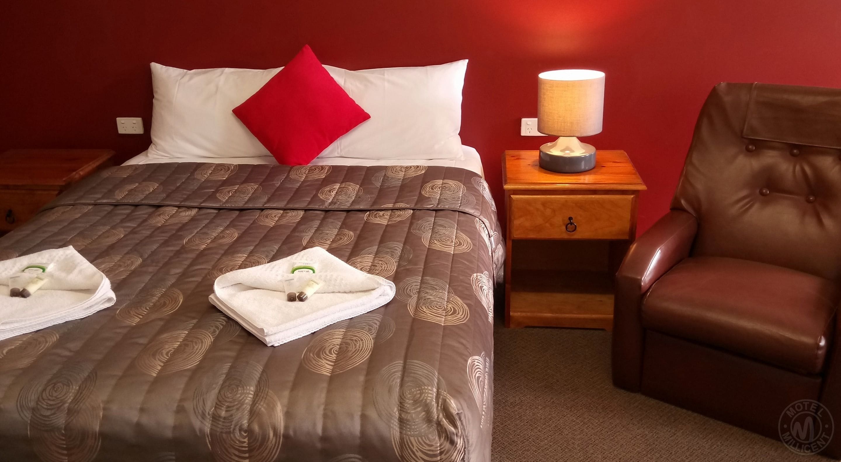 A neatly made bed with a brown patterned bedspread, a red pillow, and two towels with toiletries on the side. There is a wooden nightstand with a lamp and a brown leather chair beside the bed.