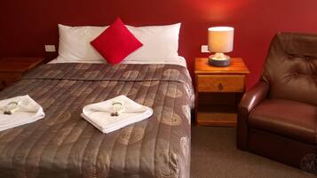 A neatly made bed with a brown patterned bedspread, a red pillow, and two towels with toiletries on the side. There is a wooden nightstand with a lamp and a brown leather chair beside the bed.