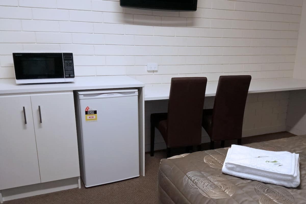 A small kitchenette with a microwave, refrigerator, and a power outlet on a white tiled wall. There are two brown chairs and a bed with a beige patterned bedspread.