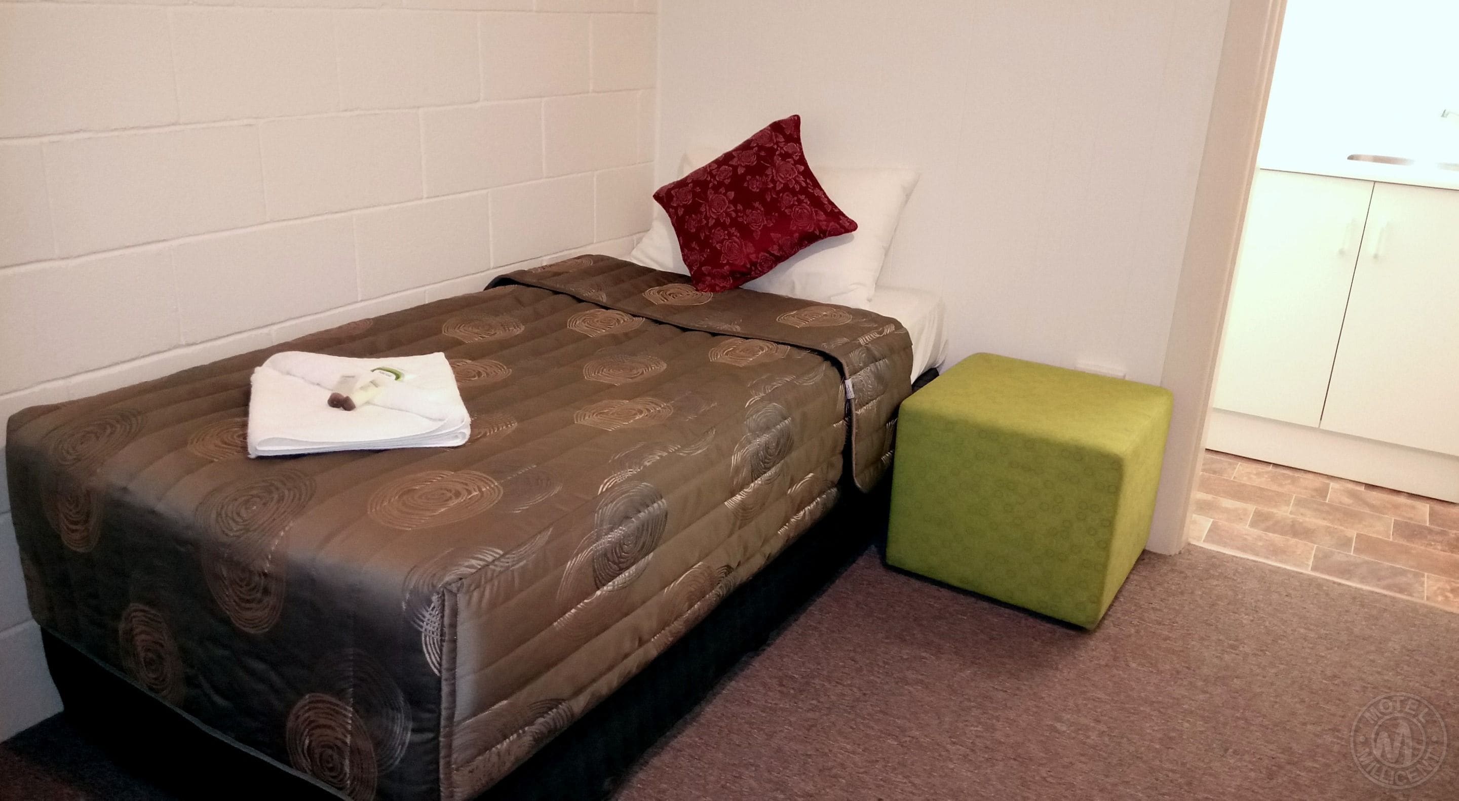 Una cama individual con una colcha marrón estampada, un cojín rojo y un reposapiés verde en una habitación con paredes blancas y piso de baldosas.