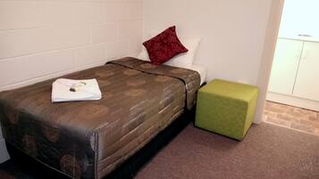Una cama individual con una colcha marrón estampada, un cojín rojo y un reposapiés verde en una habitación con paredes blancas y piso de baldosas.
