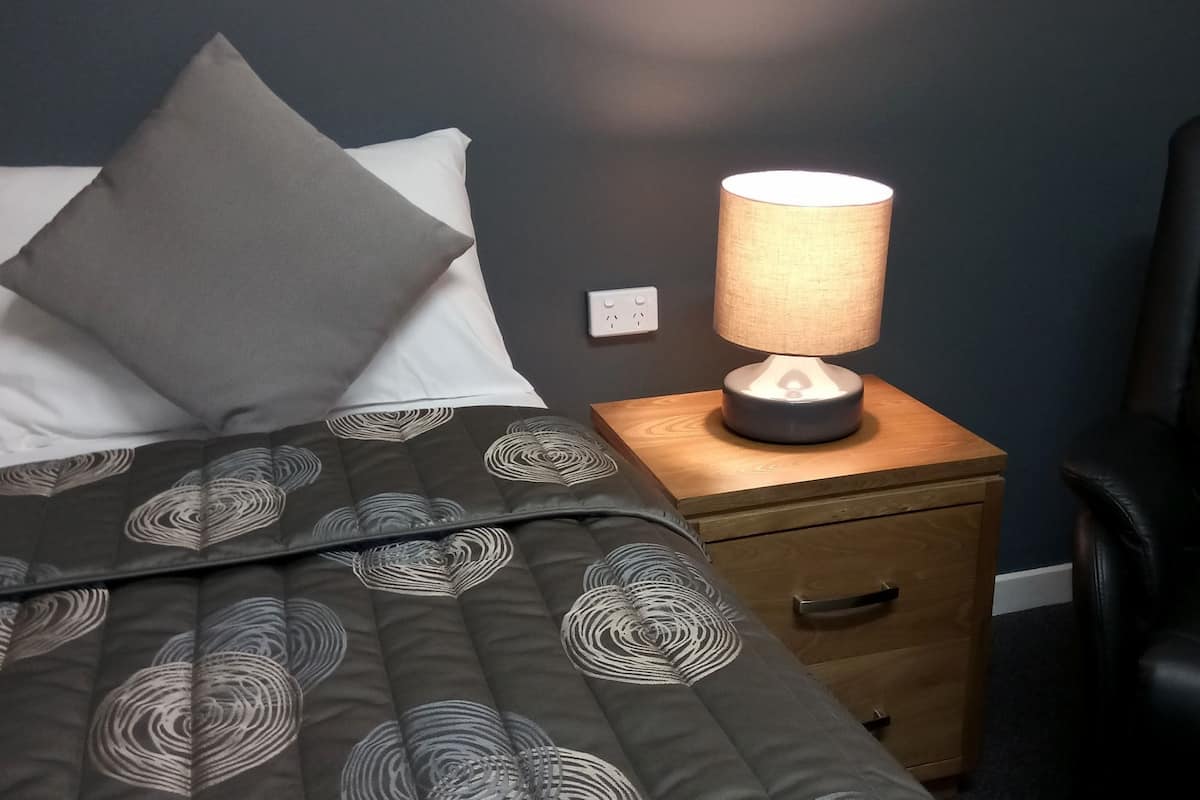 A neatly made bed with a patterned duvet, a bedside lamp, a wooden nightstand with a drawer, and a black leather chair.