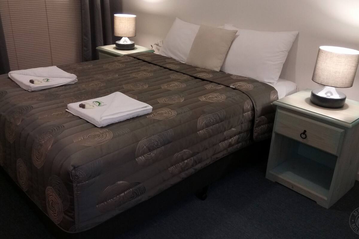 A hotel room with a large bed, two bedside tables, and a nightstand. Each table has a lamp, and the bed has two towels and a green bottle.