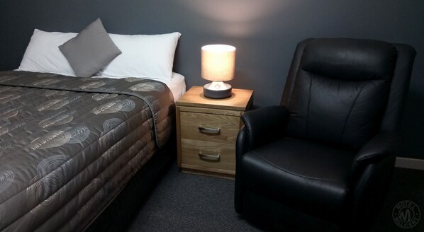 A neatly made bed with a patterned duvet, a bedside lamp, a wooden nightstand with drawers, and a black leather chair. - Millicent Motel (Millicent)