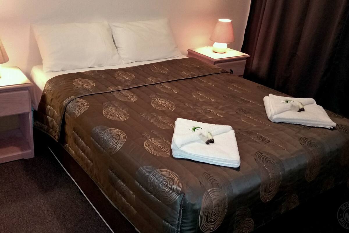 A neatly made bed with a patterned bedspread, two pillows, and two folded towels on the foot of the bed. There are two bedside tables with lamps and a small drawer.