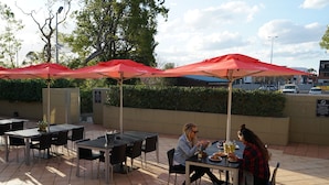 An outdoor dining area with tables and red umbrellas, surrounded by greenery and a building in the background. - Guildford Hotel (Guildford)
