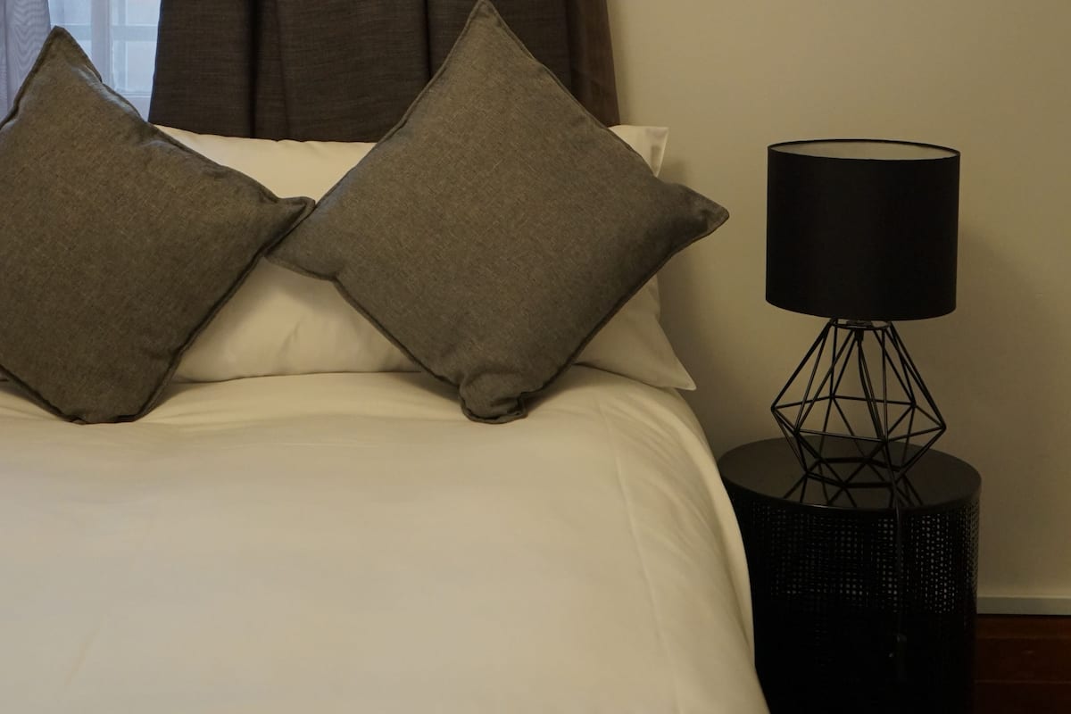 A neatly made bed with two pillows, a black lamp on a side table, and a window with curtains.