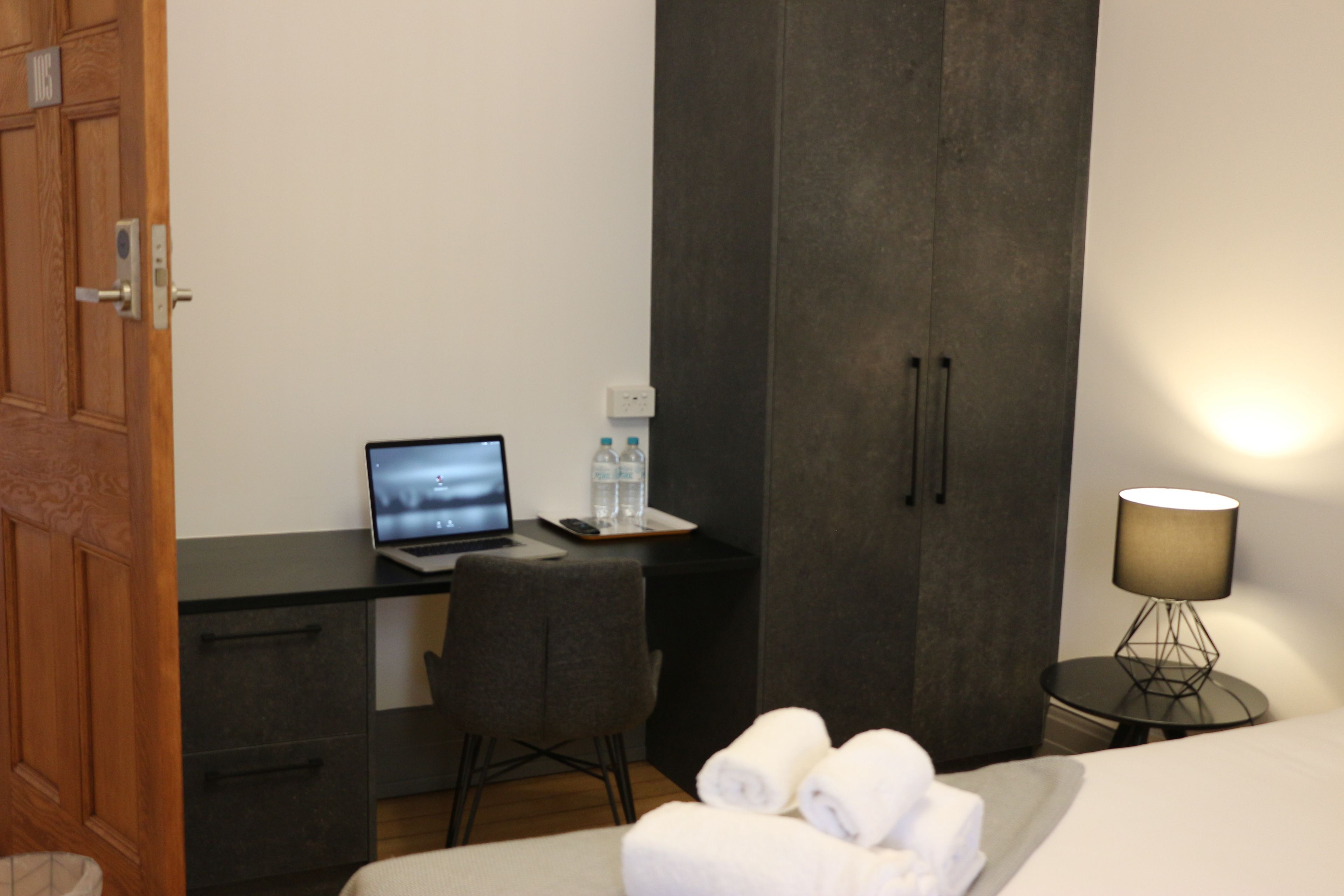 A hotel room with a wooden door, a desk with a laptop, a chair, a bed with towels, a small table with a lamp, and a wardrobe.