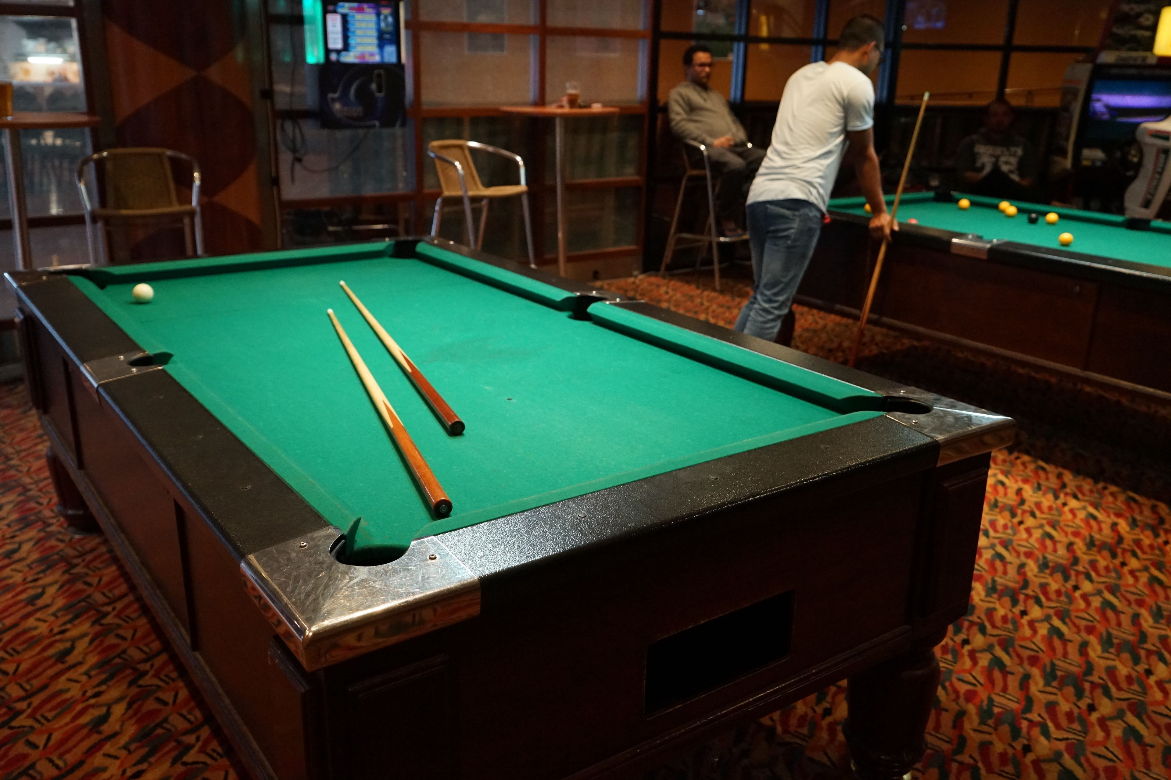 Une table de billard recouverte d’un drap vert, un queue de billard et plusieurs billes. On aperçoit des chaises et des tables à l’arrière-plan, et une personne se tient près de la table.