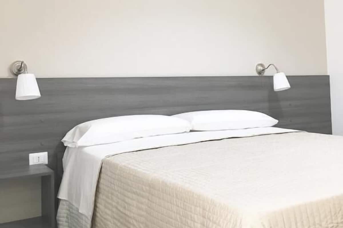 Uma cama bem arrumada com cabeceira cinza, duas luminárias de apoio e um aparelho de ar condicionado na parede.