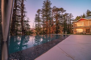 A swimming pool with a stone edge, surrounded by tall trees and a two-story building with a red roof. - Manla Homes Resort (Shimla)