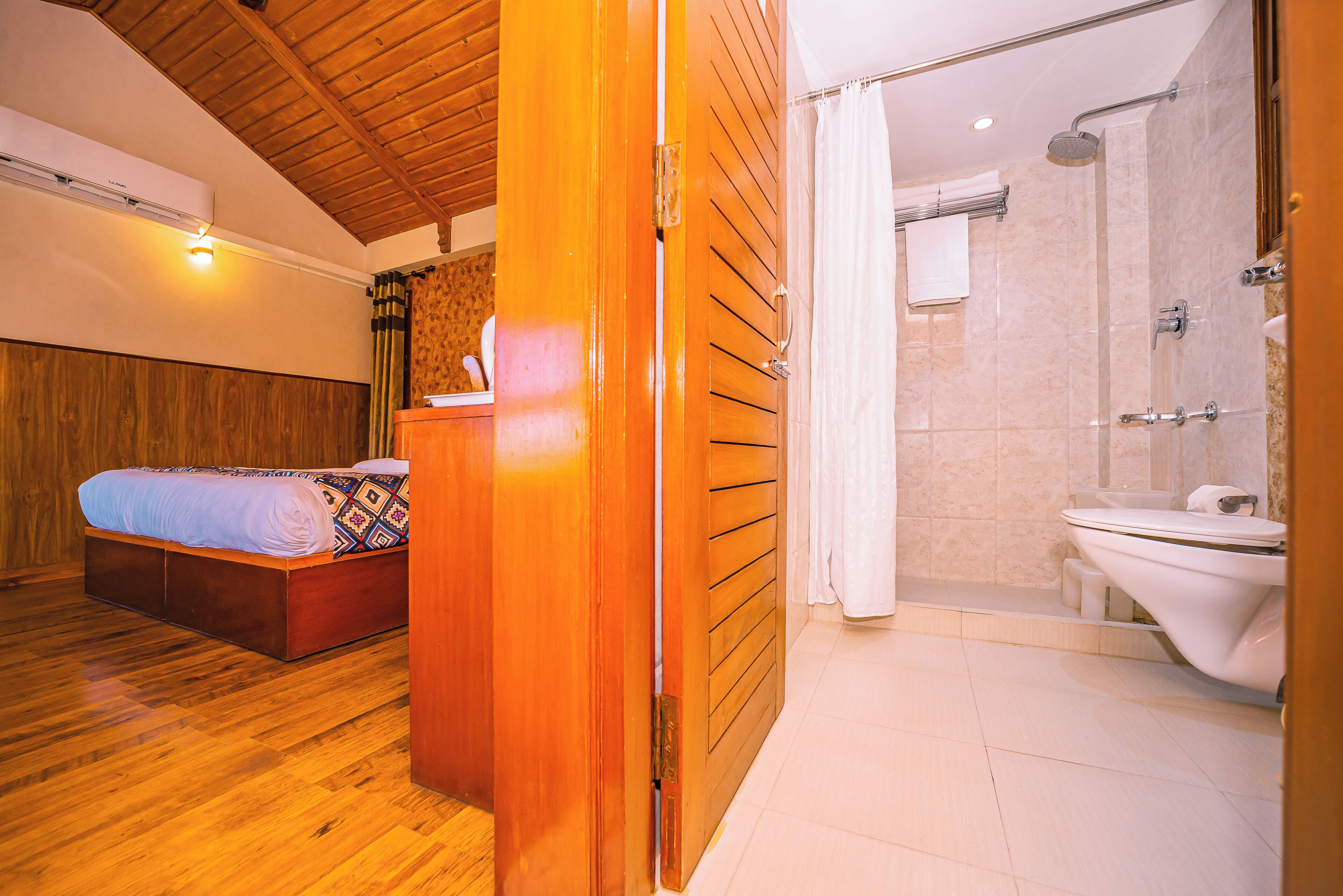 A hotel room with a wooden ceiling, a bed with a patterned bedspread, a bathroom with a shower, and a toilet.
