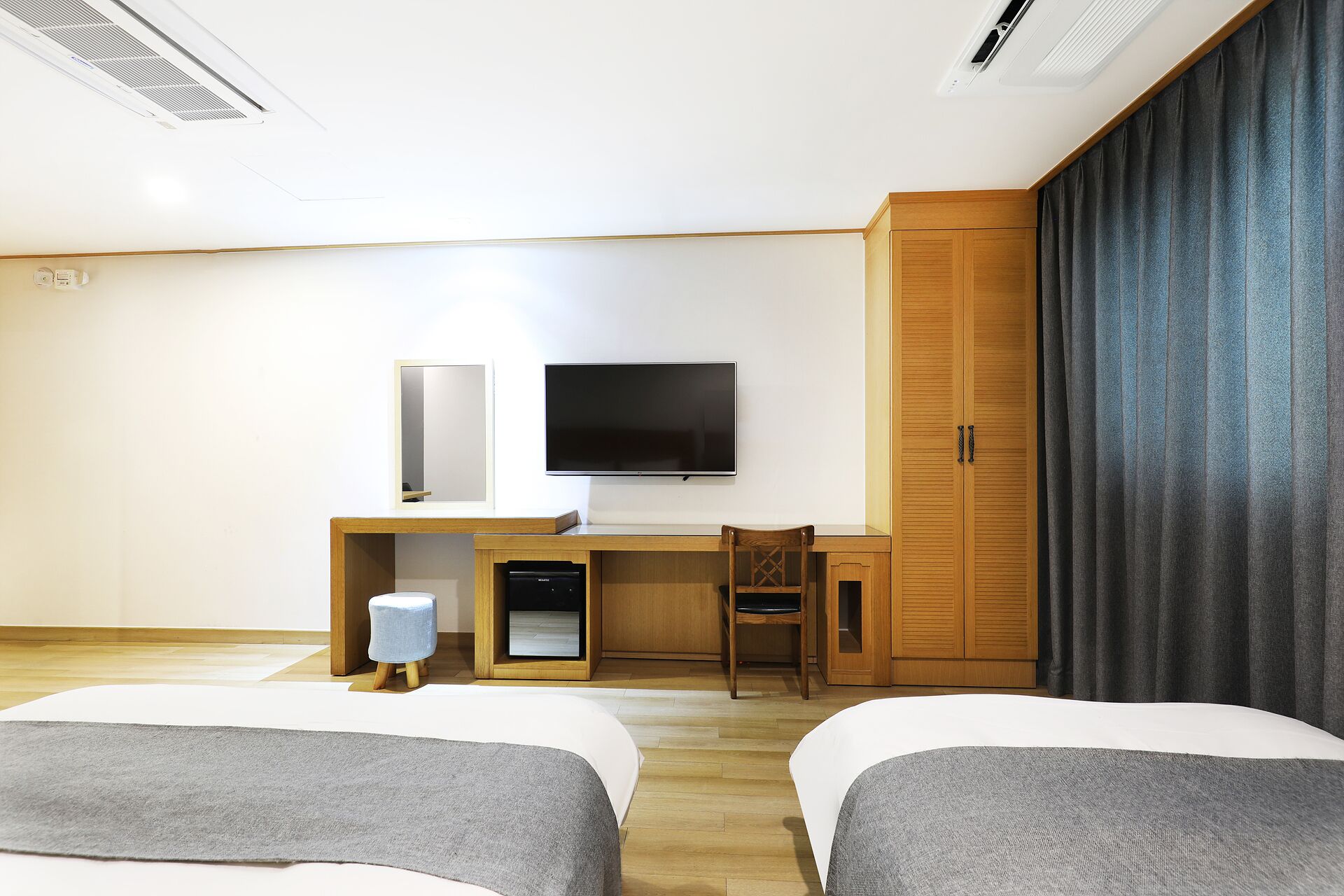 A hotel room with two beds, a desk with a TV, a wardrobe, and a small table with a lamp.