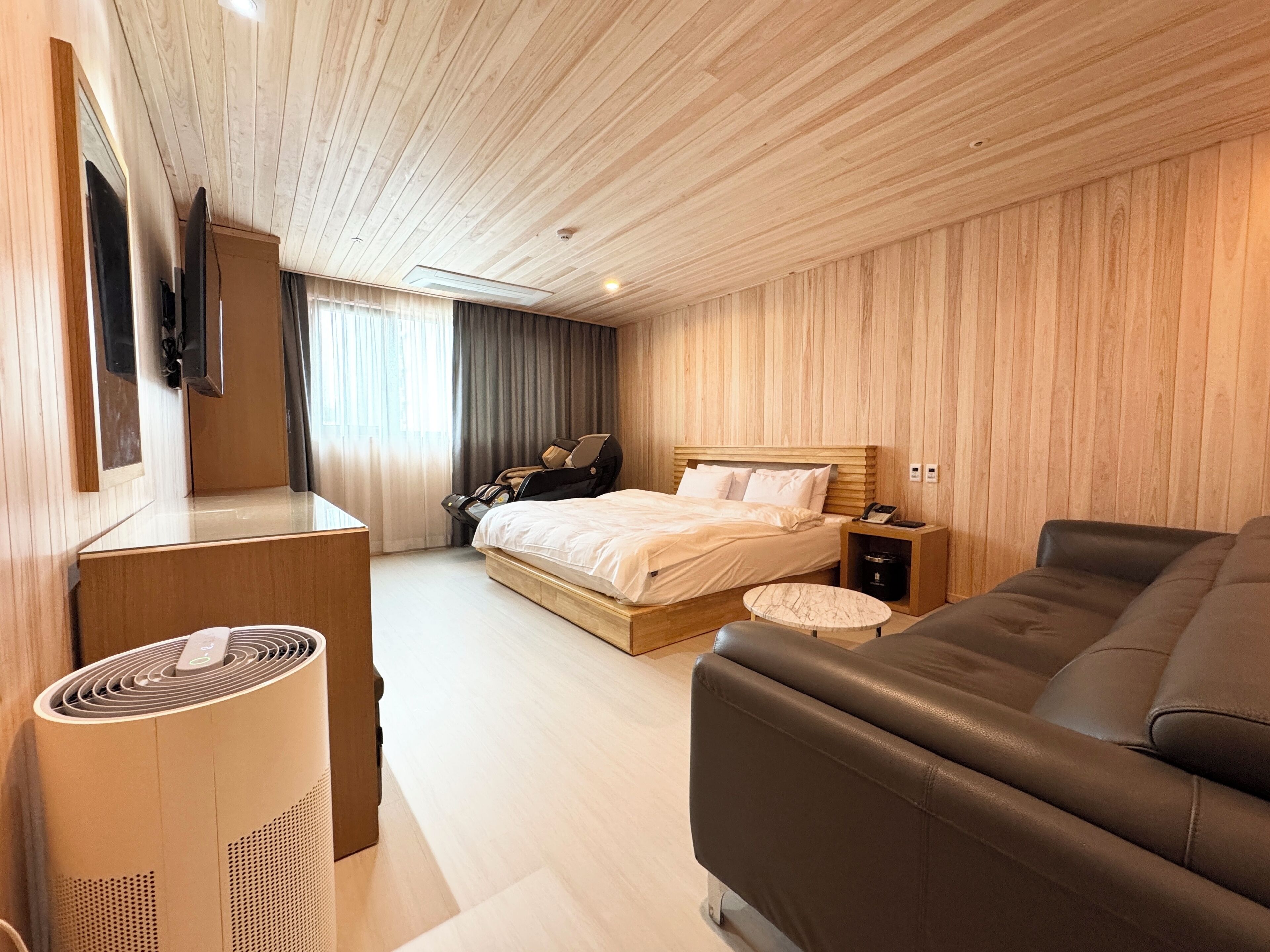 Una habitación de hotel moderna con techo de madera, cama con sábanas blancas, sofá de cuero, sillón de masaje y televisor de pantalla plana montado en la pared.