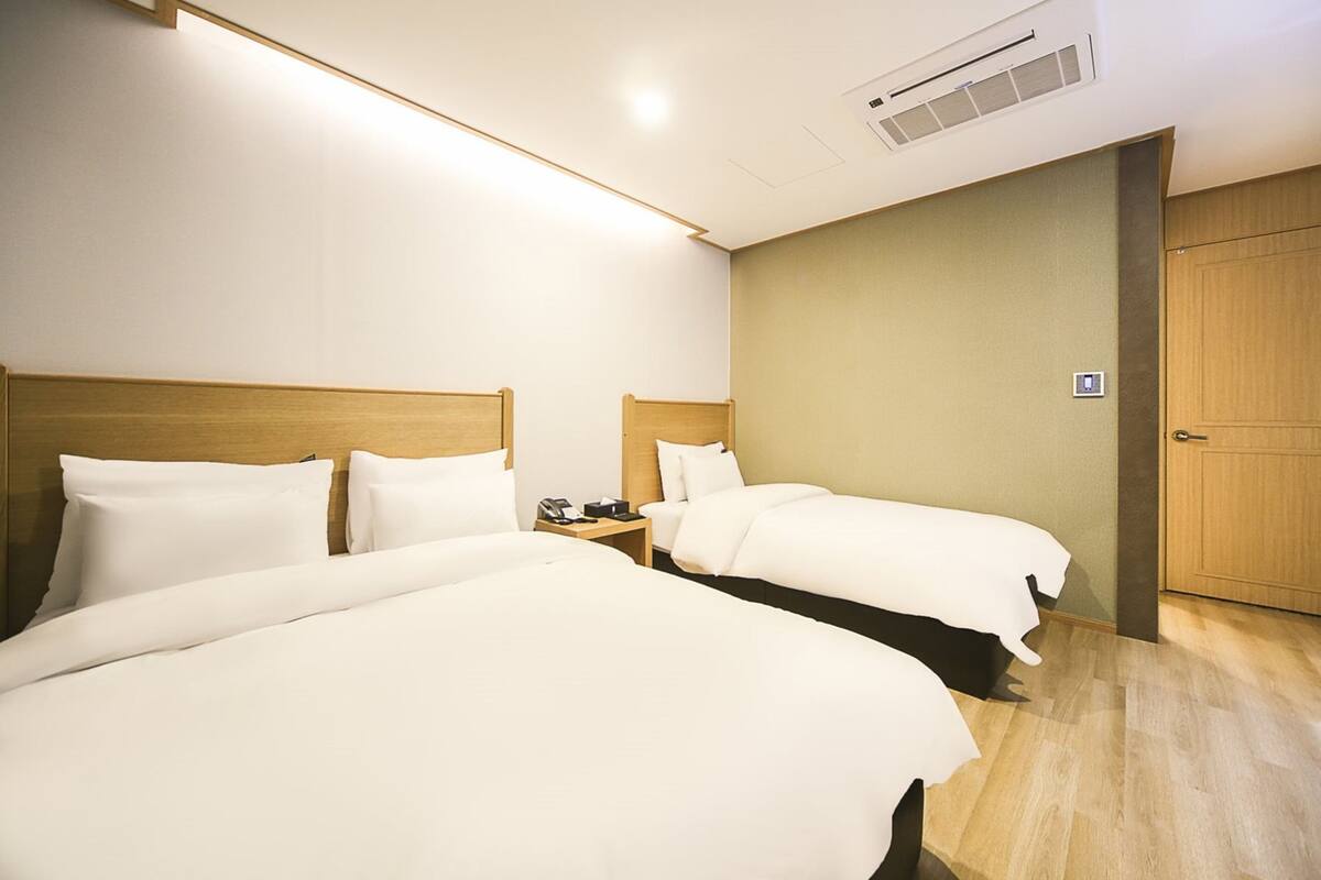 A hotel room with two beds, a wooden headboard, a nightstand, a wall-mounted air conditioner, and a wooden door.