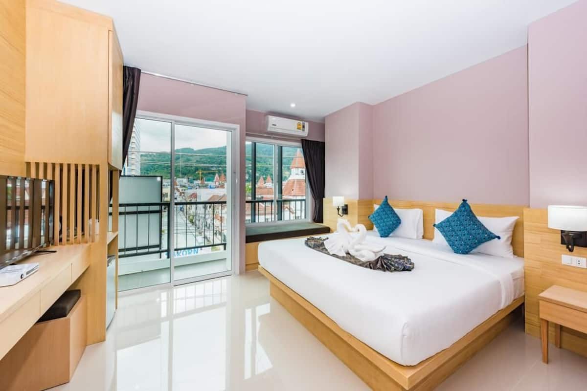 A modern hotel room with a large bed, a desk, a TV, and a balcony with a view of buildings.