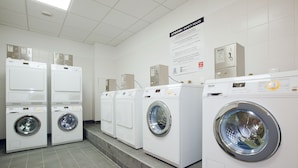 Une buanderie équipée de plusieurs machines à laver et de plusieurs séchoirs, un panneau indiquant les consignes de sécurité et un système de ventilation fixé au mur.