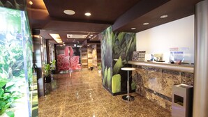 Uma área de recepção de hotel com piso de mármore, balcão de atendimento, um vaso de plantas e um grande cartaz de uma flor.
