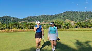 Zwei Personen gehen auf einem Golfplatz, im Hintergrund eine hügelige Landschaft.