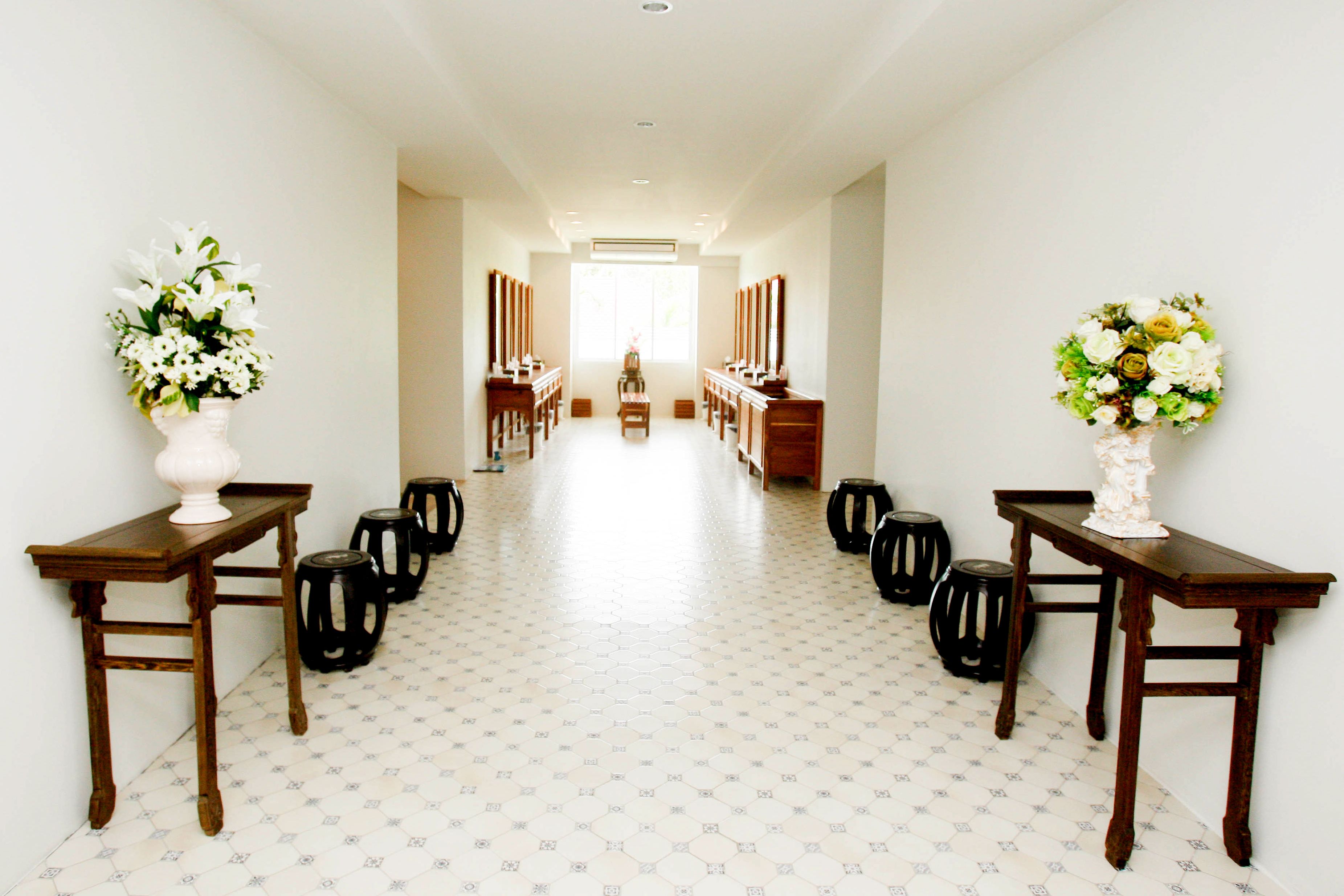 Un pasillo de hotel con piso de baldosas, mesas de madera con jarrones de flores, y vista al área de las habitaciones en la distancia.