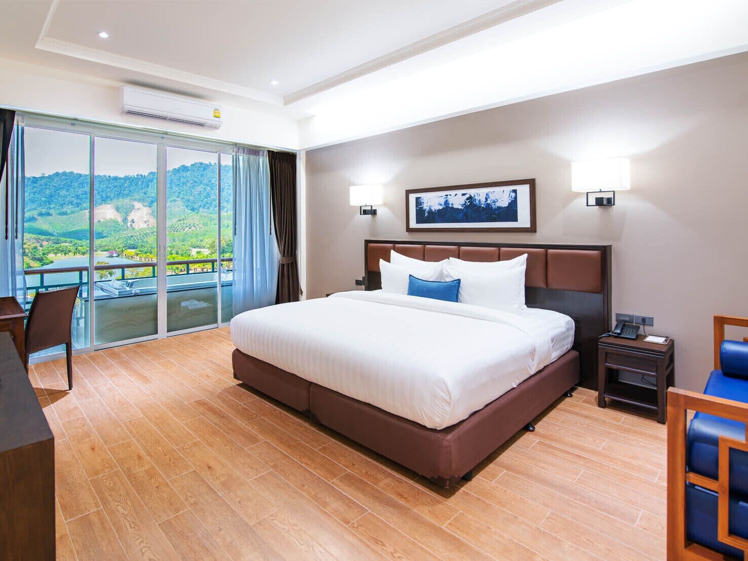 A hotel room with a large bed, a desk, and a chair. There is a balcony with a view of the mountains.