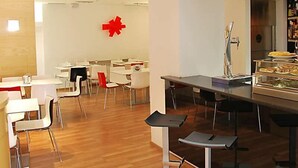 Un área de comedor moderna con mesas y sillas blancas, un elemento decorativo rojo en el centro y un mostrador de buffet con diferentes opciones de comida.