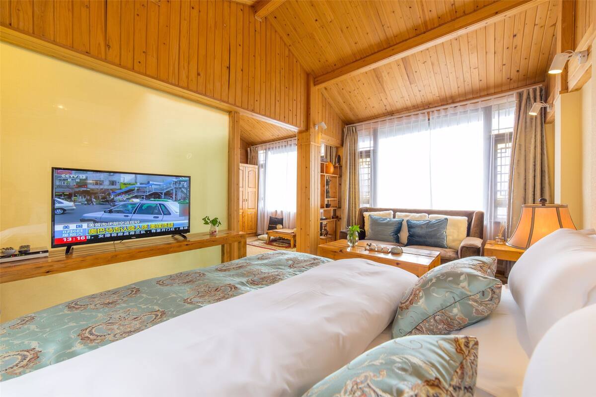 Habitación de hotel con techo de madera, televisor montado en la pared, sofá, cama con ropa de cama estampada y ventana con cortinas.