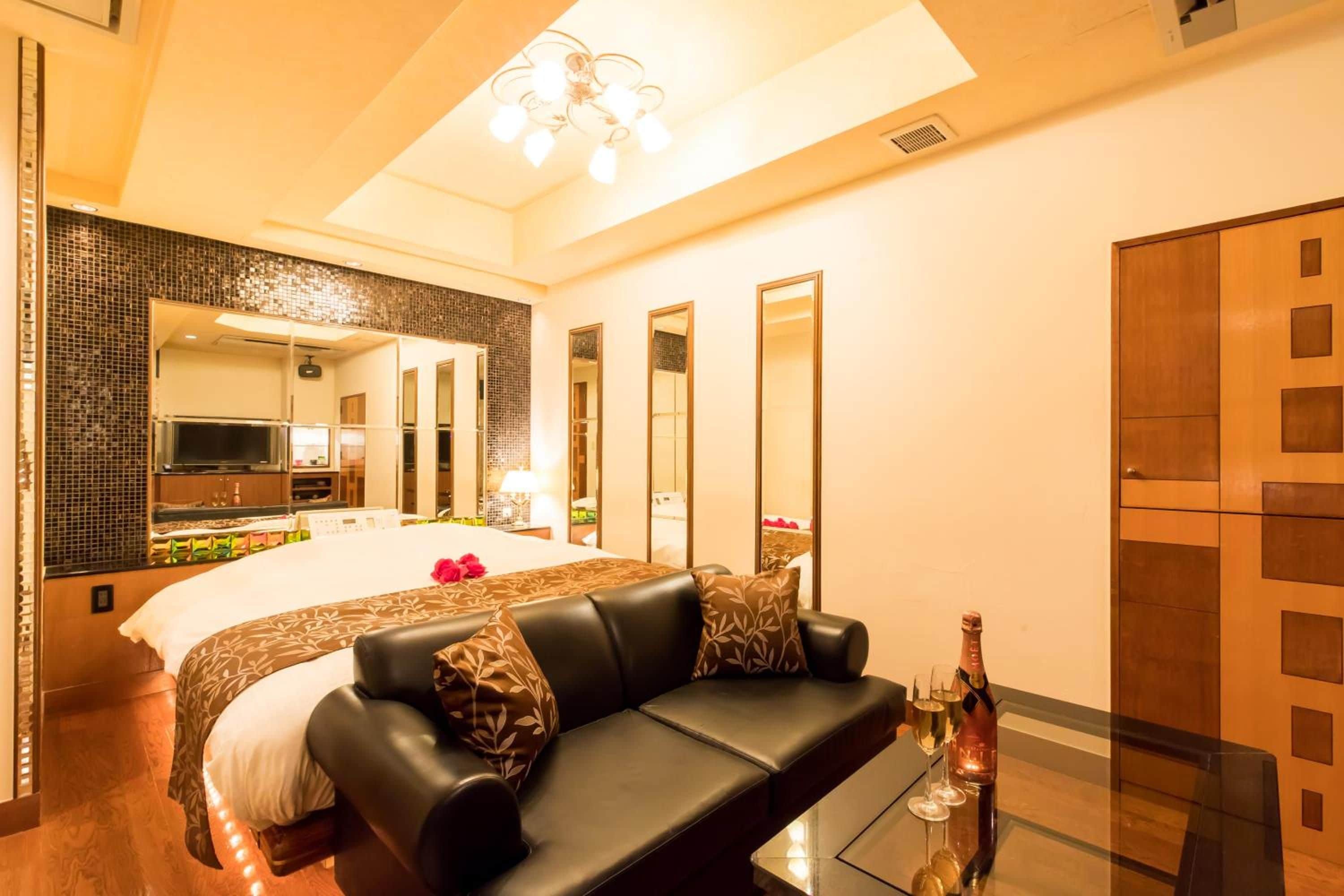 A modern hotel room with a large bed, a leather sofa, a glass coffee table, and a minibar.