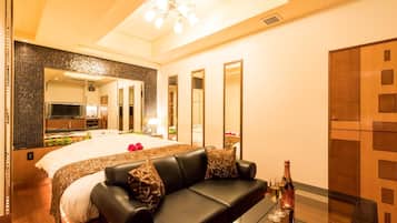 A modern hotel room with a large bed, a leather sofa, a glass coffee table, and a minibar.