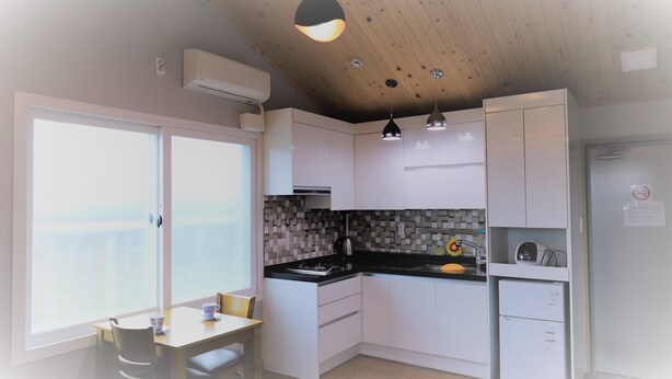 A modern kitchen with white cabinets, a black countertop, and a built-in stove. There is a dining area with a table and chairs, and a window providing a view. - Woojinsam Pension (Jeju City)