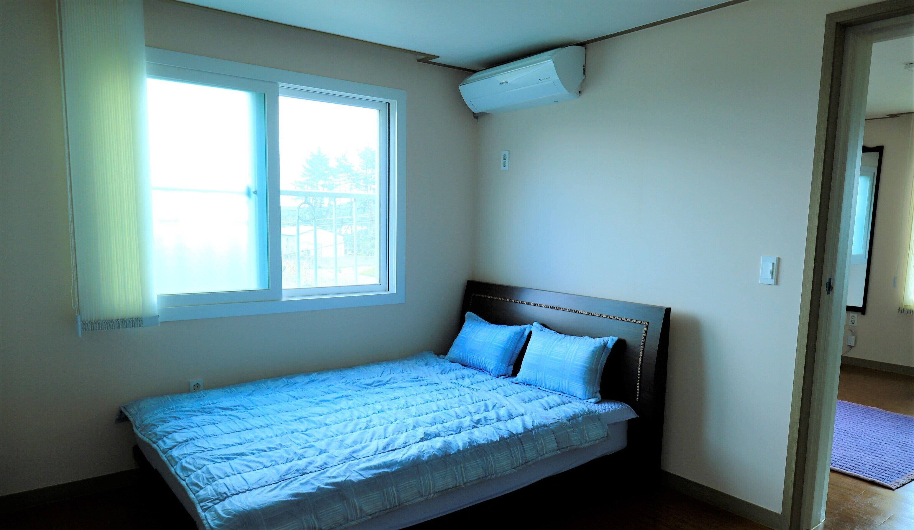 A single bed with a headboard, a window with blinds, and an air conditioning unit on the wall.
