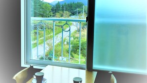A room with a window overlooking a balcony with a metal railing and a view of greenery and hills. - Woojinsam Pension (Jeju City)