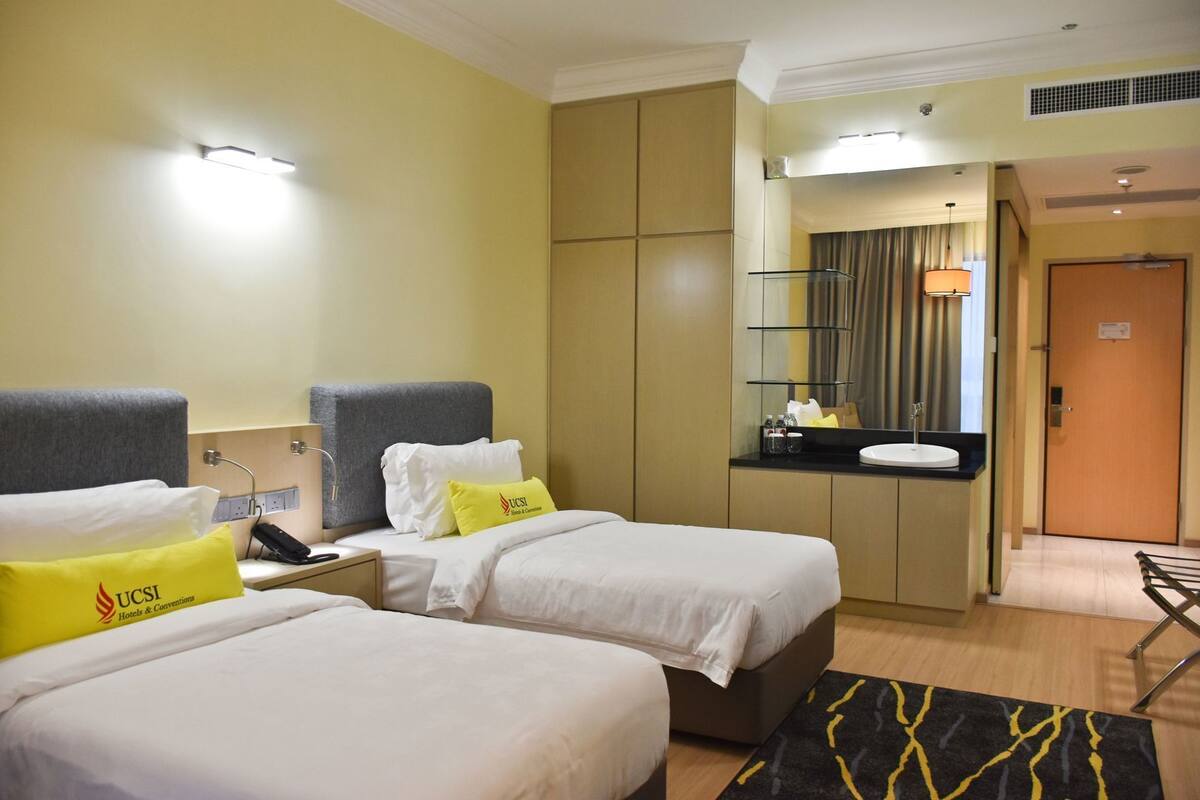 A hotel room with two beds, a small desk, a mirror, and a bathroom visible in the background.