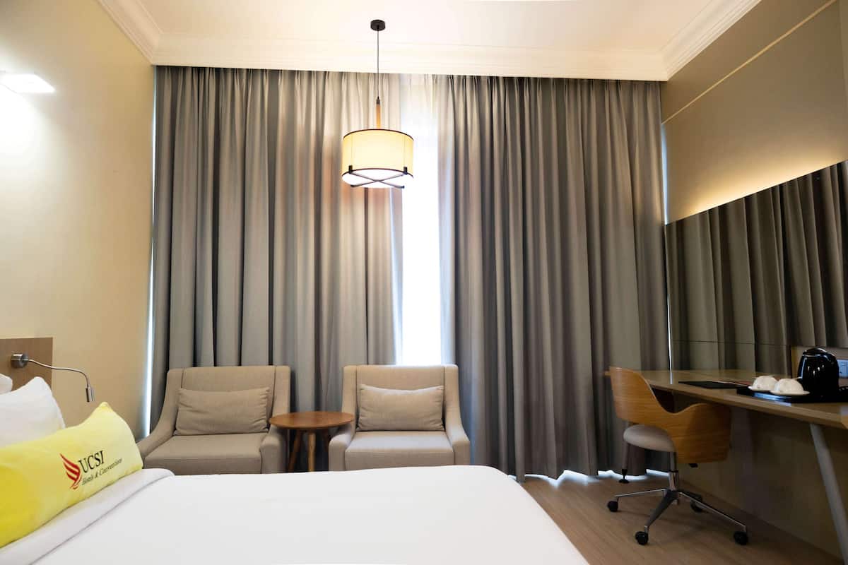 A hotel room with a bed, two armchairs, a desk, a lamp, and a large window with curtains.