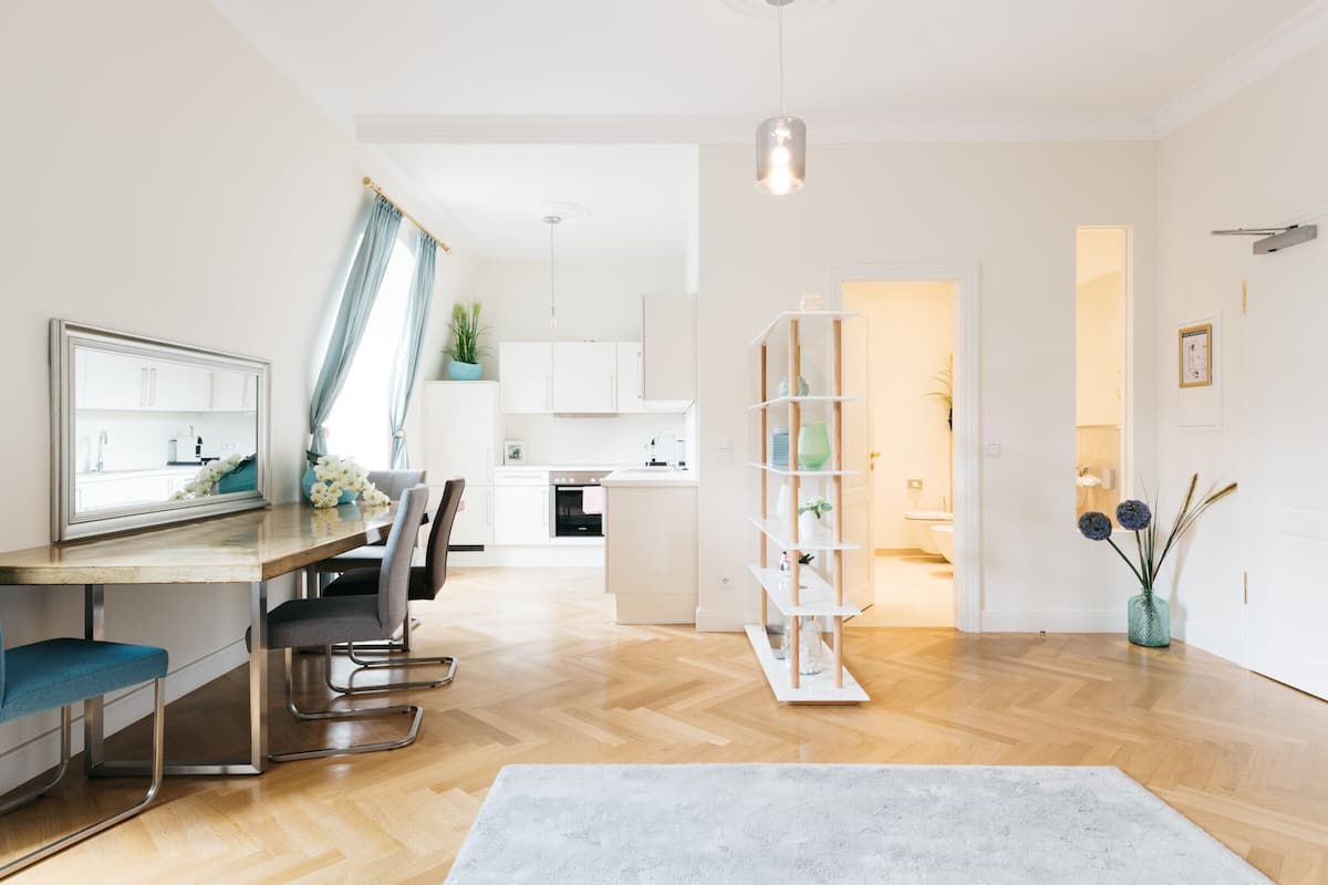 A modern kitchen with white cabinets, a dining area with a wooden table and chairs, and a hallway leading to a bedroom.