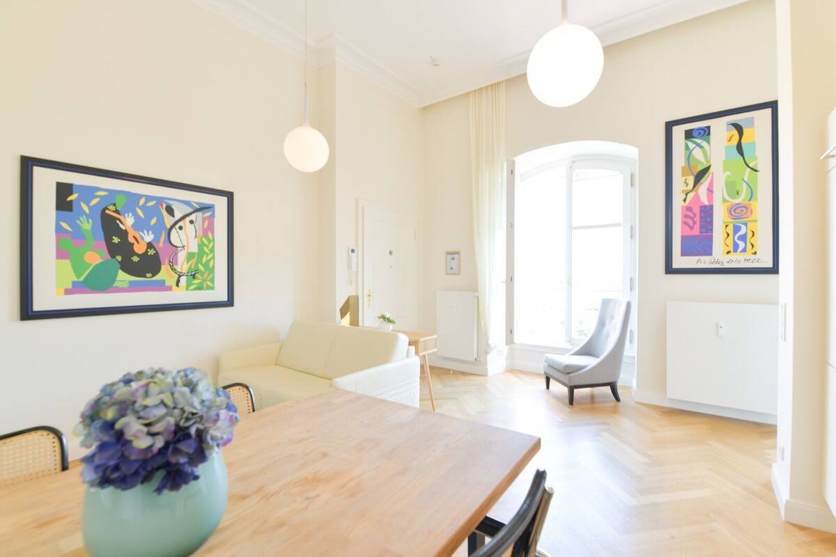 A spacious living room with a wooden dining table, a sofa, and a grey armchair. There are two large windows providing natural light and a view of the outdoors.