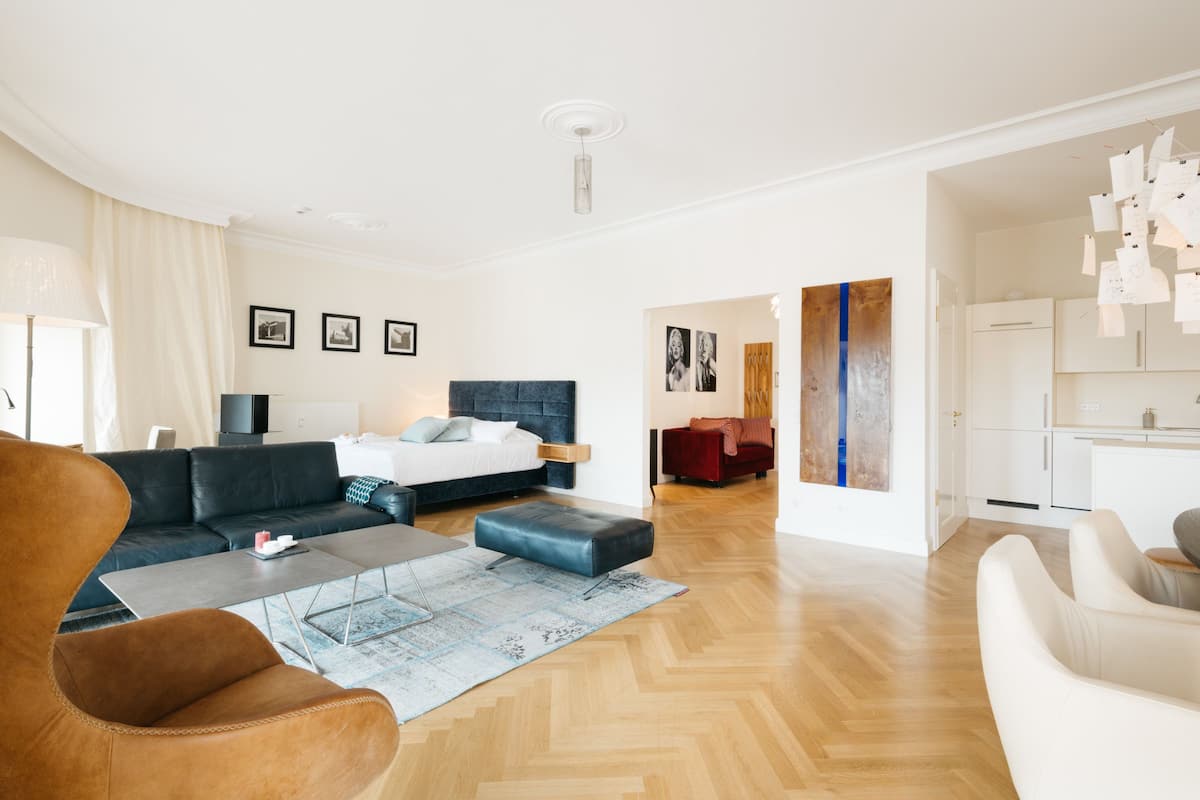A modern living room with a sofa, coffee table, and ottoman. There is a bed with a headboard and a red chair in the background.