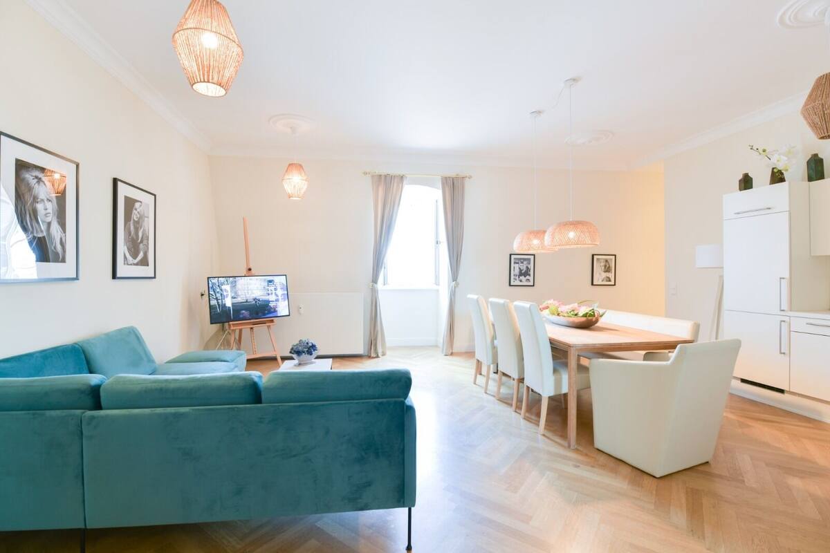 A modern living room with a blue sofa, a dining table with chairs, a TV on a stand, and a kitchen area with white cabinets.