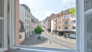 Une vue sur la rue, prise depuis une fenêtre, avec des bâtiments des deux côtés, un arbre au centre et un ciel dégagé.