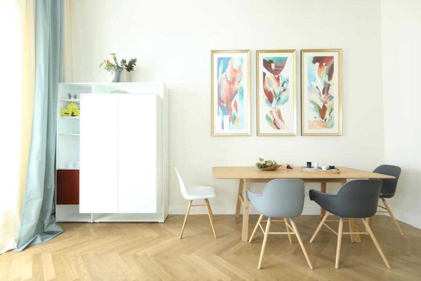 A modern dining area with a wooden table, four chairs, and three framed abstract paintings on the wall.