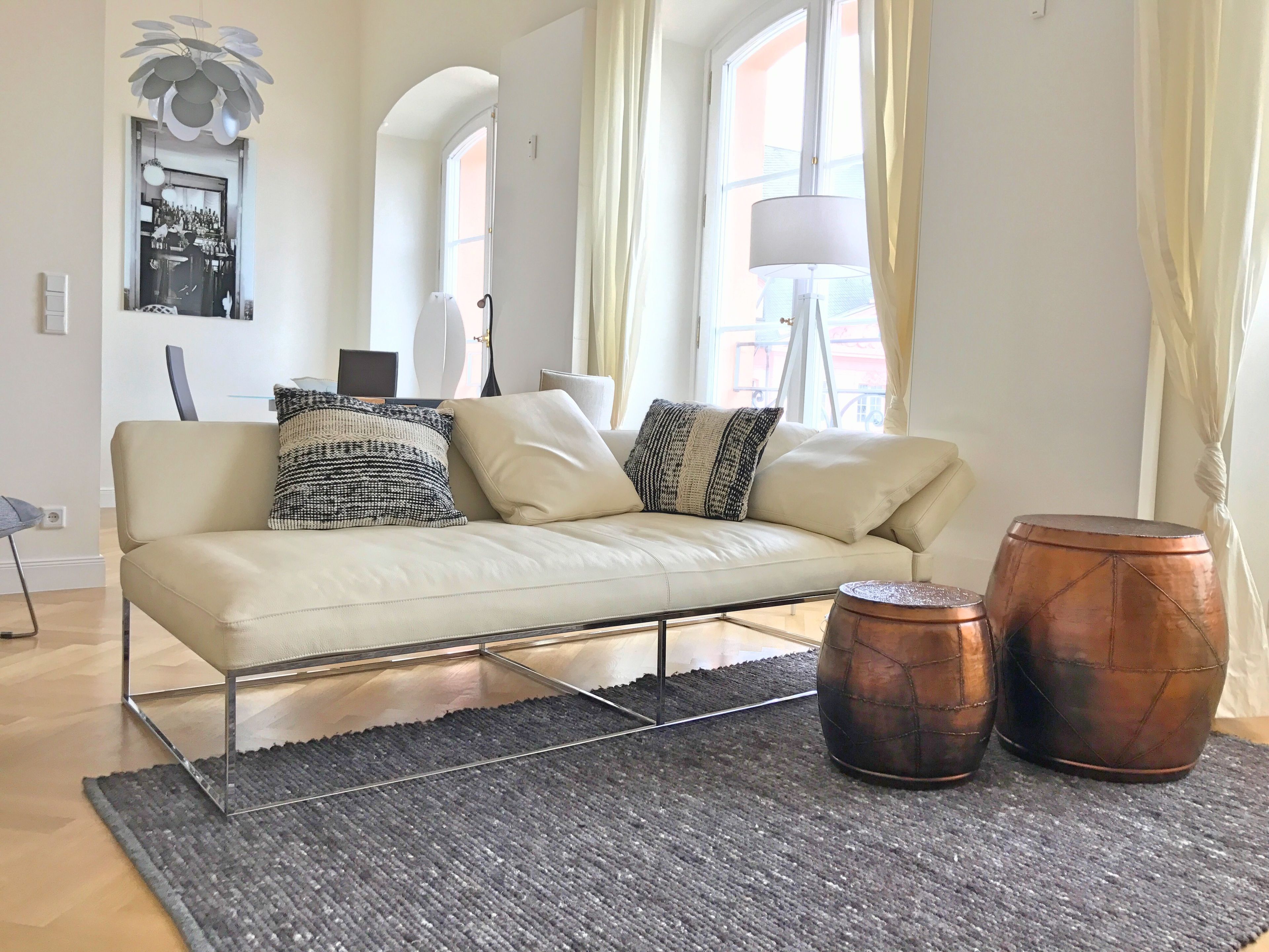 A modern living room with a sofa, two round copper side tables, a grey rug, and a large window with curtains.