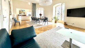 A modern living room with a sofa, dining table, and chairs. There is a TV mounted on the wall and a kitchen in the background. - The Apartment Suite Osteiner Hof (Mainz)