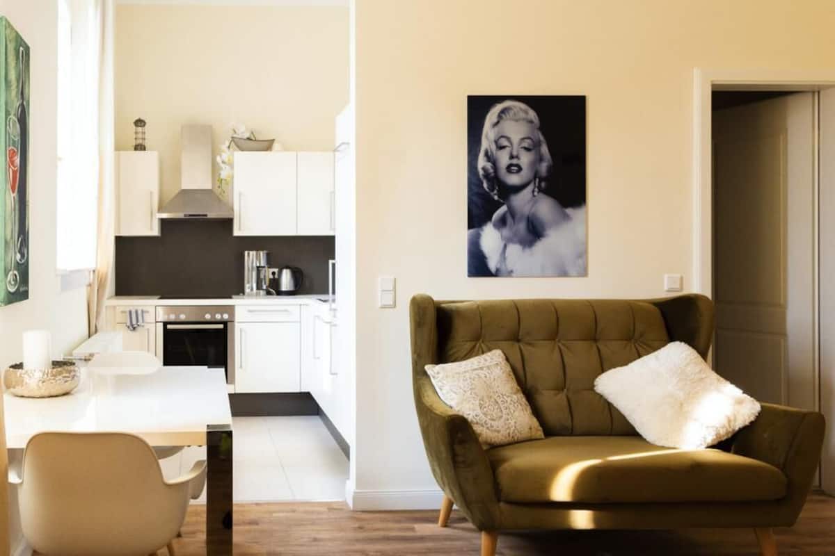 A modern living room with a green sofa, a dining area with a white table, and a kitchen in the background.