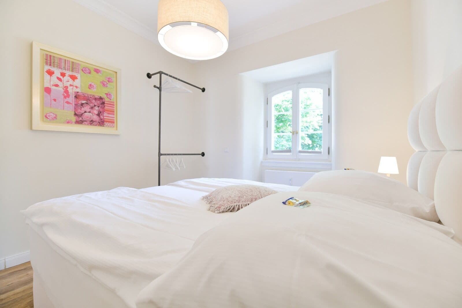 A bedroom with a bed, a hanging light fixture, a window, and a wall-mounted artwork.