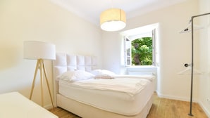 A modern bedroom with a large bed, a floor lamp, and a view of greenery outside. - The Apartment Suite Osteiner Hof (Mainz)