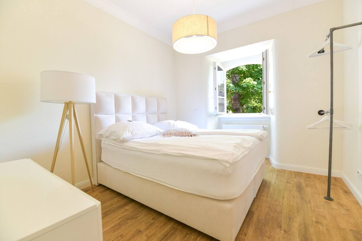 A modern bedroom with a large bed, a floor lamp, and a view of greenery outside.