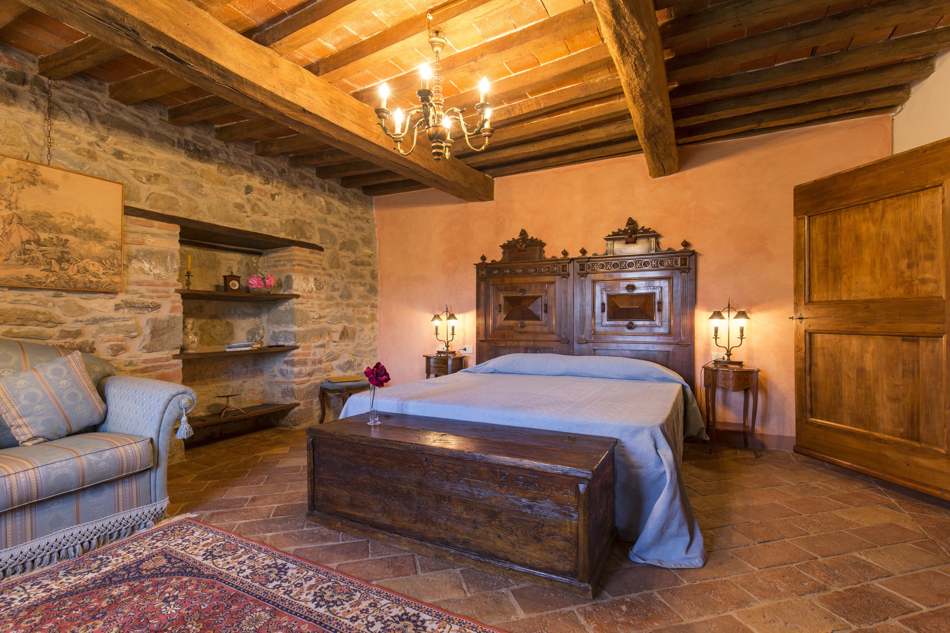 A cozy bedroom with a stone wall, a wooden bed, a nightstand, and a chandelier.